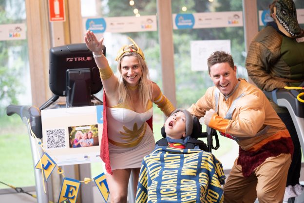 A young boy is in his wheelchair, with a blanket over his body. A man and a woman are on either side, posing in superhero costumes.