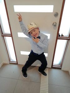 A child is dancing and posing for the camera, he is wearing a boy tie and fedora hat.