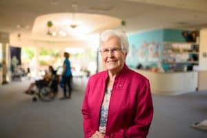 Sister Margaret is standing in the children's hospice smiling at the camera. There are nurses and children in the background.
