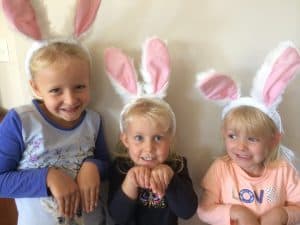 Three young girls are smiling at the camera, they are wearing bunny rabbit ears at Easter time.