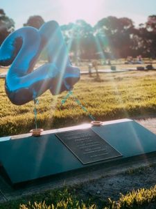 An image of the sun shining on a gravestone, with balloons attached of a 2 and a 1 for his 21st birthday