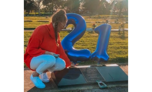 A lady is kneeling by a gravestone, there are balloons tied to it of a 2 and a 1 for his 21st birthday. The sun is shining.
