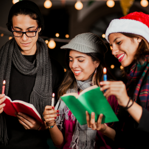 Three young adults are holding carol books and candles. One is wearing a Santa hat.