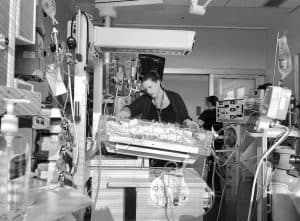 A woman is standing in a hospital room, watching over her baby in a crib. There are many machines and wires visible.