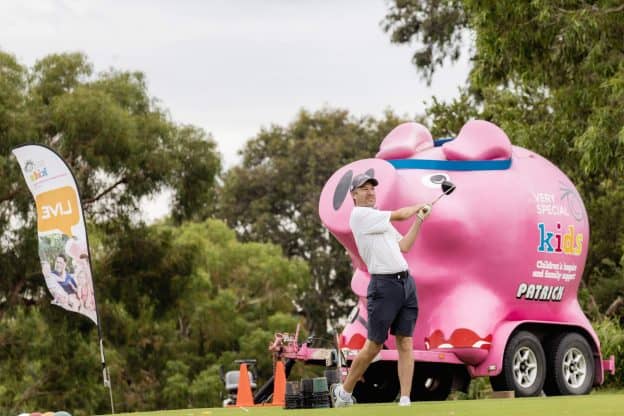 A man is playing golf outdoors. Behind him is a giant pig shaped trailer.