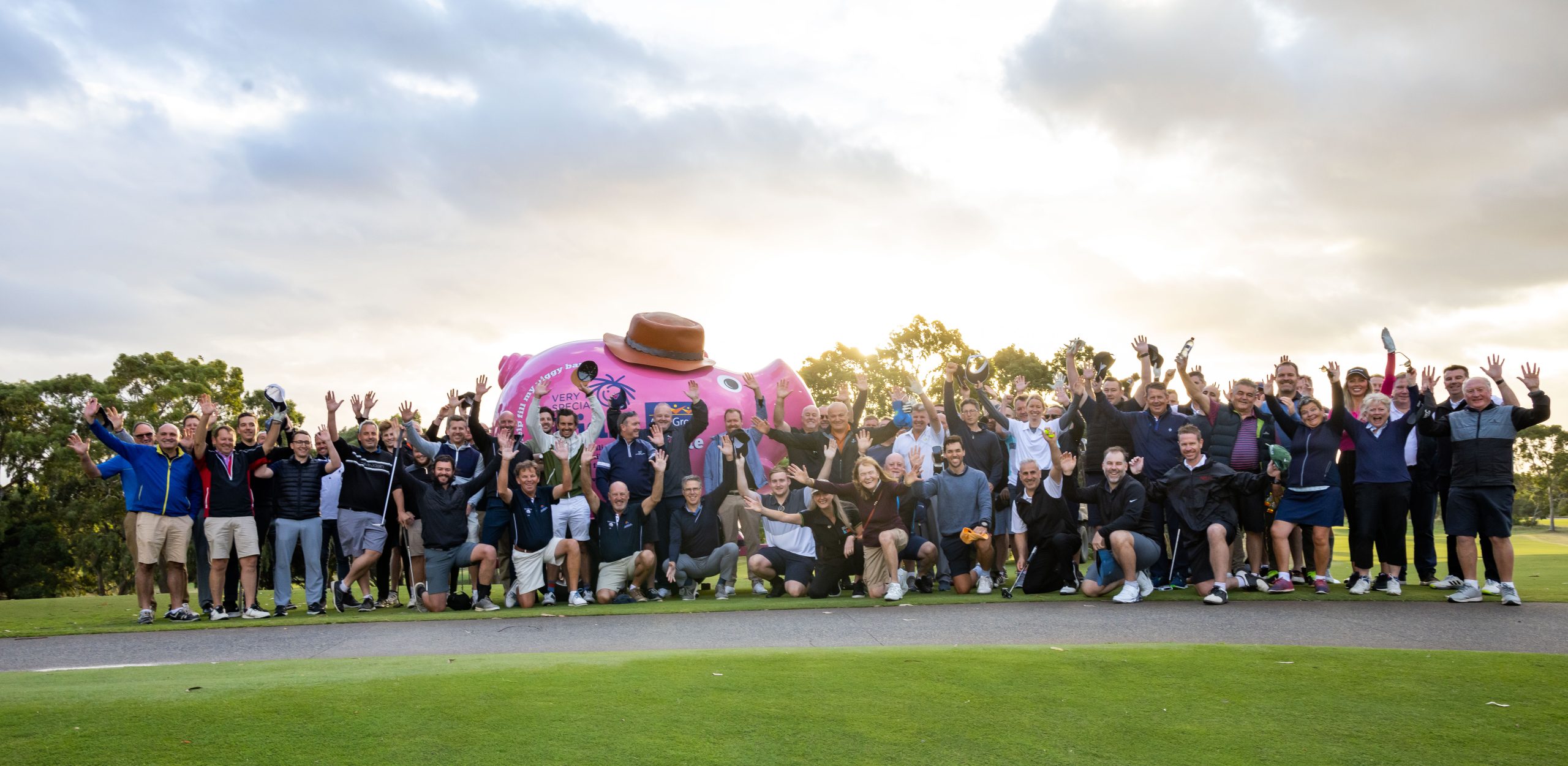 A large group of people are standing outdoors, in front of a giant piggy bank, with the sunrise behind. They have their arms in the air and are smiling at the camera.