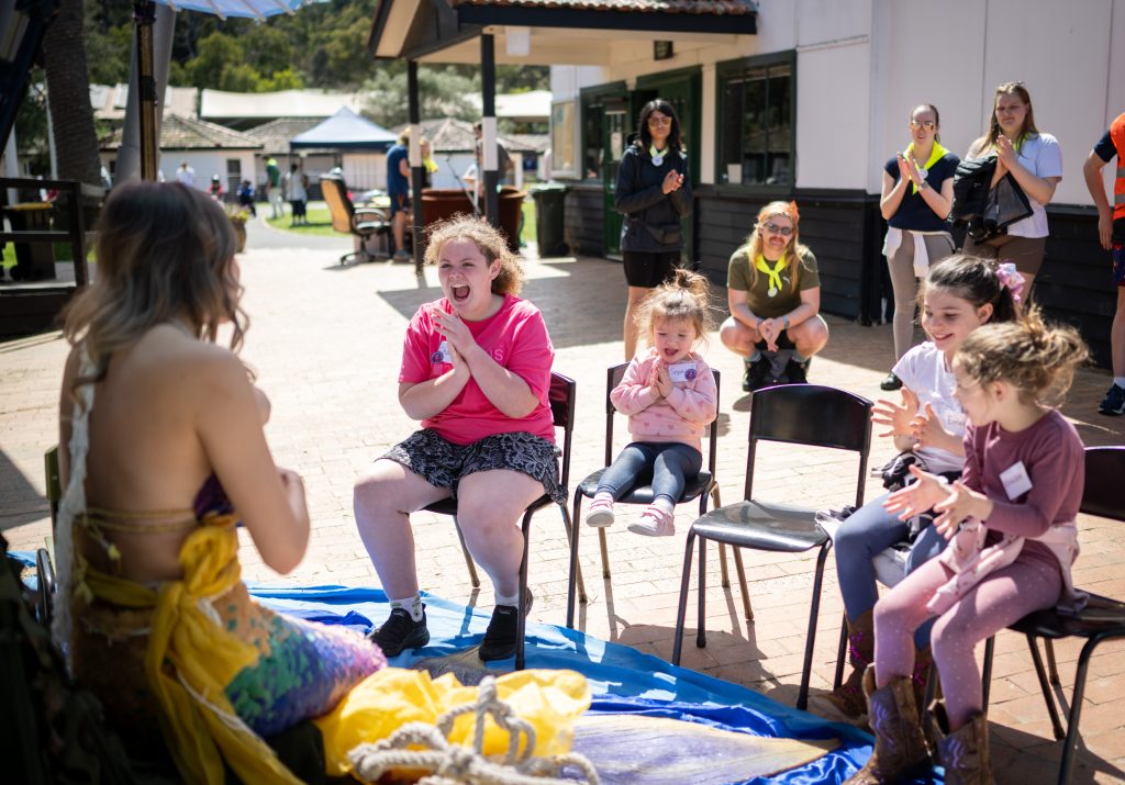 A group of children and adolescents are watching a woman dressed as a mermaid as she reads to them. They are smiling and clapping