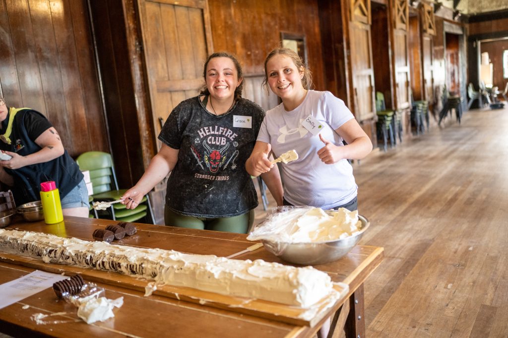 Two young women are smiling at the camera as they decorate a long cake inside. 