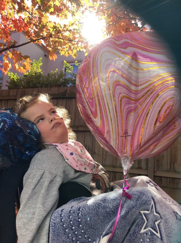 A young girl is sitting in her wheelchair outdoors. She has a pink coloured balloon and the sun is shining through the trees.