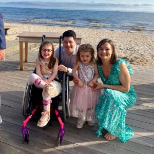 A family of two adults and two young girls are on a path in front of a beach. One of the girls is in a wheelchair and they are all smiling at the camera.