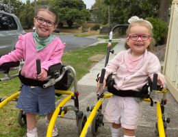Two young girls are outdoors walking with the help of walkers. They are smiling at the camera.
