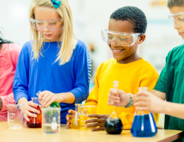 Three children are wearing safety glasses as they pour coloured liquid between test tubes and science equipment.
