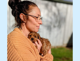 A woman is standing outdoors holding a small puppy. She is wearing glasses and facing off camera.