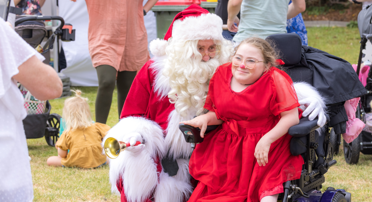 A girl is sitting in her wheelchair smiling as Santa Claus kneels next to her.